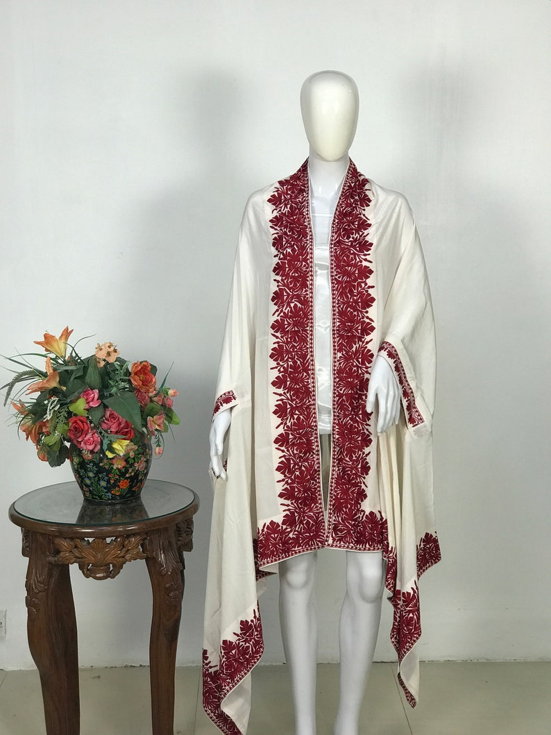 May include: A white shawl with a red embroidered floral design around the edges. The shawl is draped over a white mannequin.
