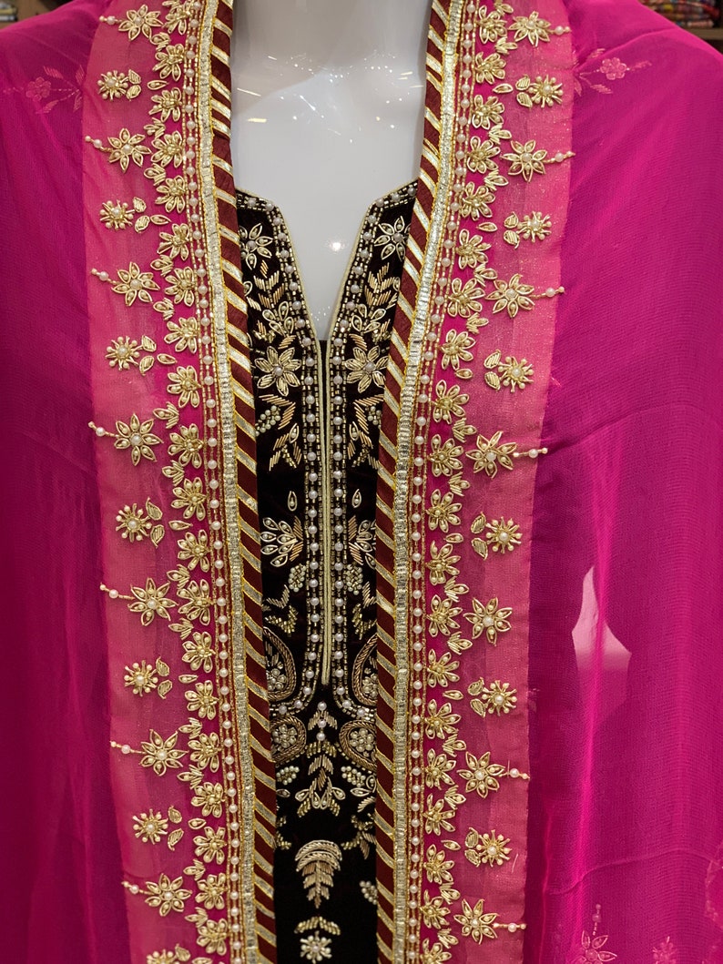 May include: A black velvet dress with gold and white embroidery. The dress has a V-neckline and is paired with a pink chiffon dupatta.