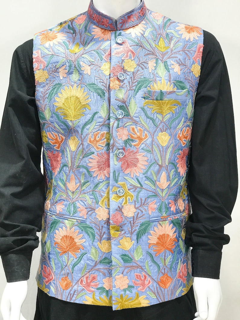 May include: A blue embroidered vest with a floral pattern in shades of pink, orange, yellow, and green. The vest has a mandarin collar and a button closure.