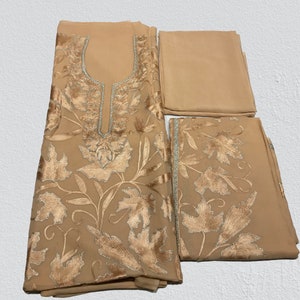 May include: A beige three-piece garment set featuring a tunic, trousers, and a scarf. The tunic has a U-shaped neckline with silver trim and intricate gold leaf embroidery. The trousers feature matching gold leaf embroidery along the hem.