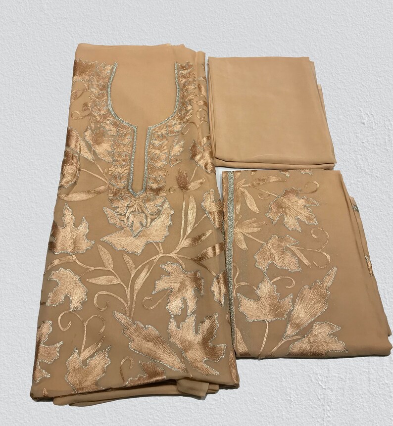 May include: A beige three-piece garment set featuring a tunic, trousers, and a scarf. The tunic has a U-shaped neckline with silver trim and intricate gold leaf embroidery. The trousers feature matching gold leaf embroidery along the hem.