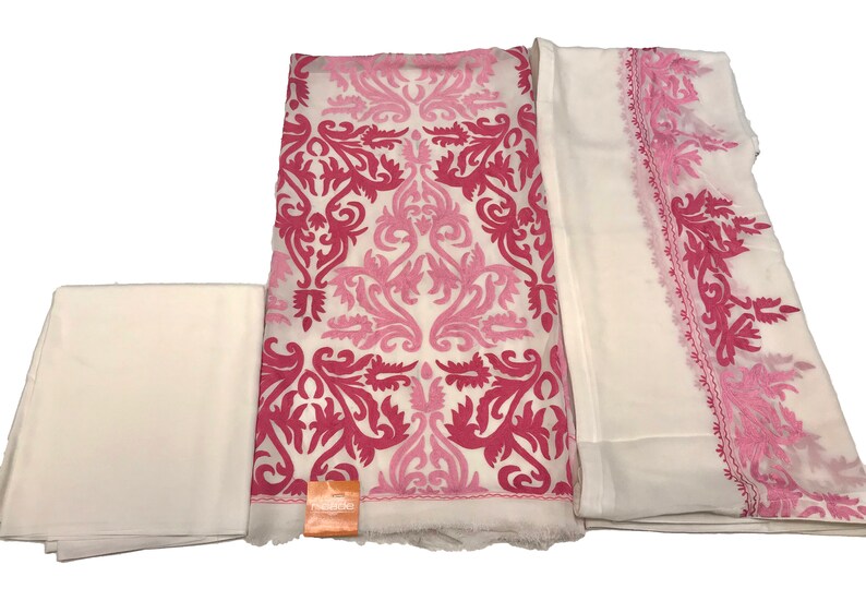 May include: A three-piece fabric set featuring a white base with vibrant pink and light pink damask-style embroidery. The set includes a plain white fabric piece, a large central piece with dense floral patterns, and a sheer white dupatta with pink borders.