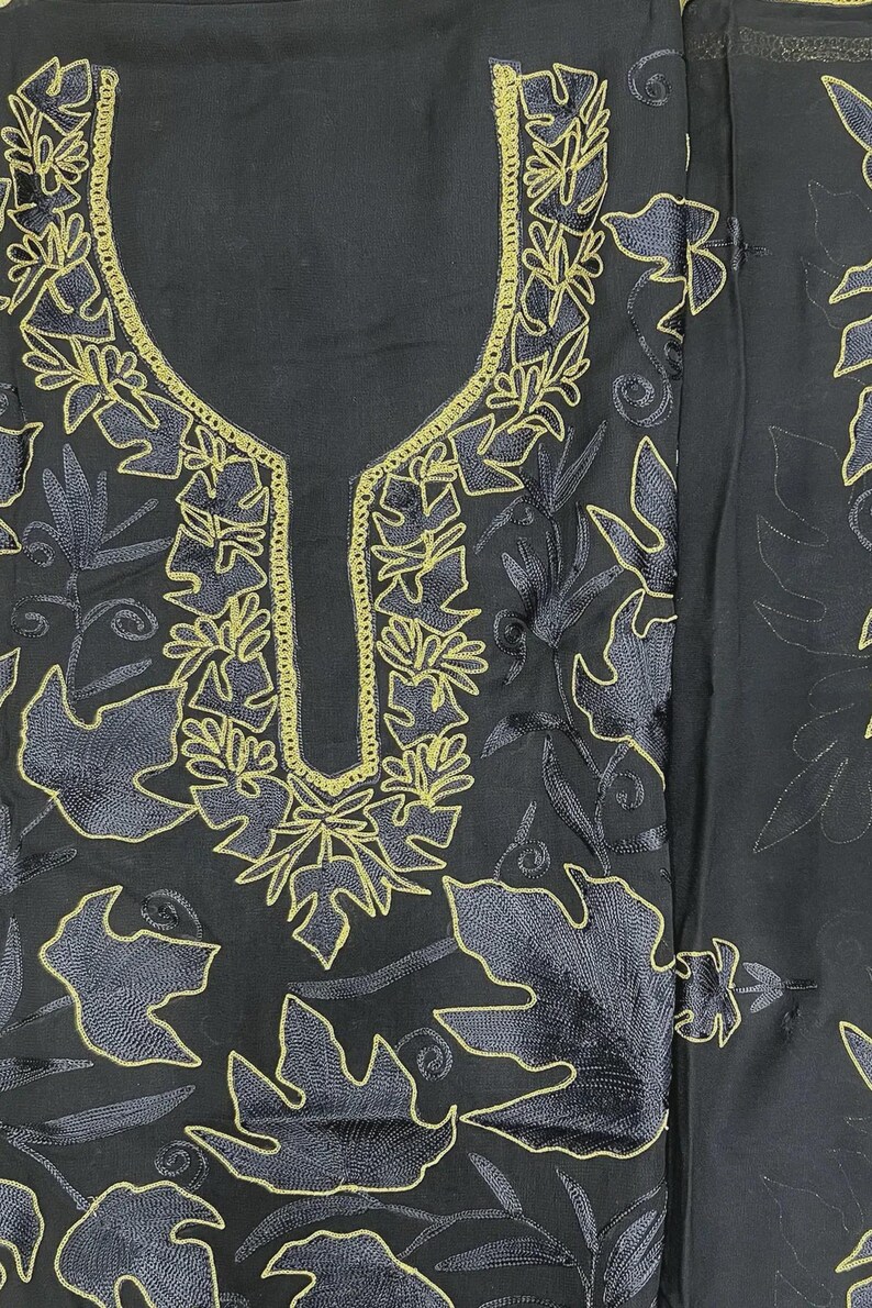 May include: A black fabric garment featuring a U-shaped neckline and intricate embroidery. The design consists of golden-yellow thread outlining large, stylized leaf patterns and swirling vine motifs that cover the front of the piece.