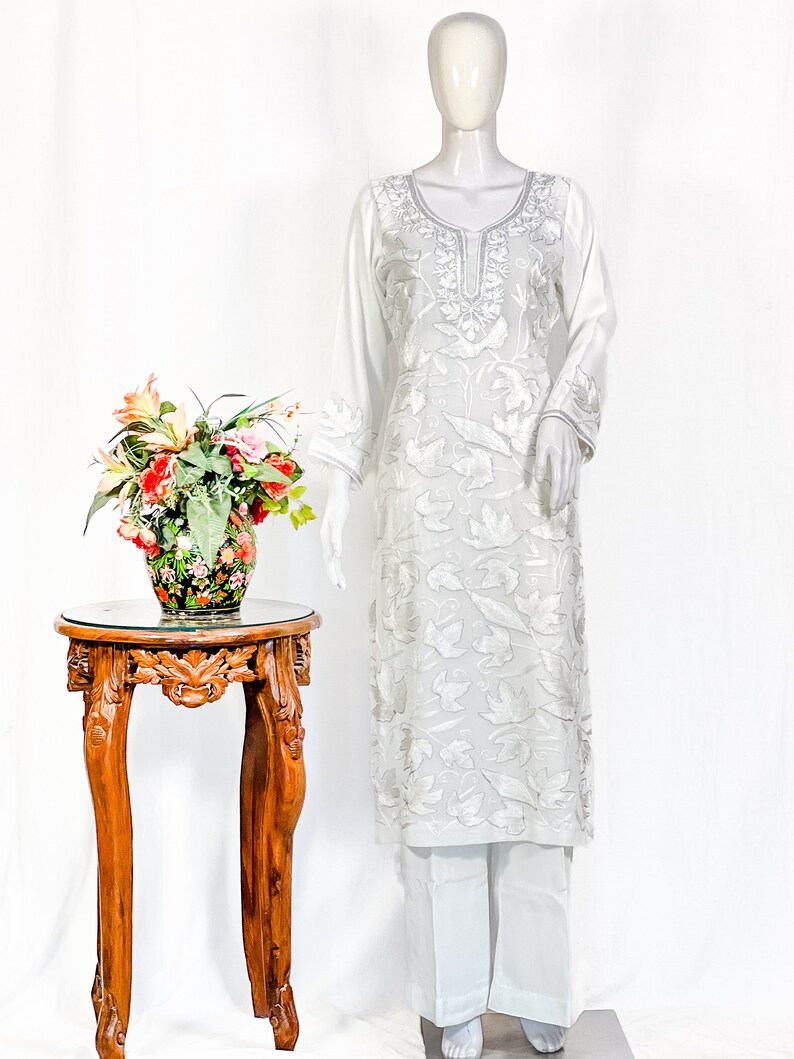 May include: A white long-sleeved tunic featuring intricate tonal floral embroidery throughout the fabric, paired with matching white straight-leg trousers. The tunic has a round neckline with detailed silver-toned stitching along the collar.