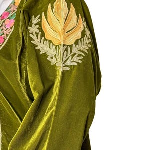May include: An olive green velvet garment featuring an embroidered golden yellow floral motif on the shoulder. The neckline displays a decorative border with pink, orange, and green floral embroidery on a cream-colored fabric base.