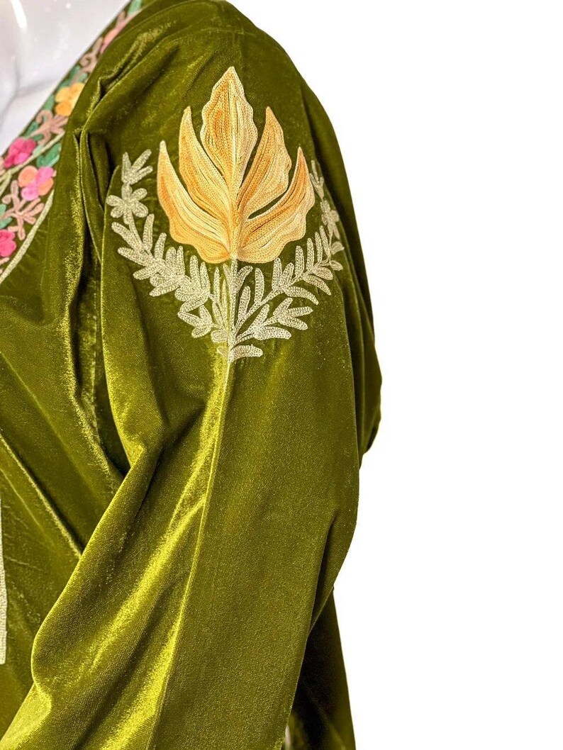 May include: An olive green velvet garment featuring an embroidered golden yellow floral motif on the shoulder. The neckline displays a decorative border with pink, orange, and green floral embroidery on a cream-colored fabric base.