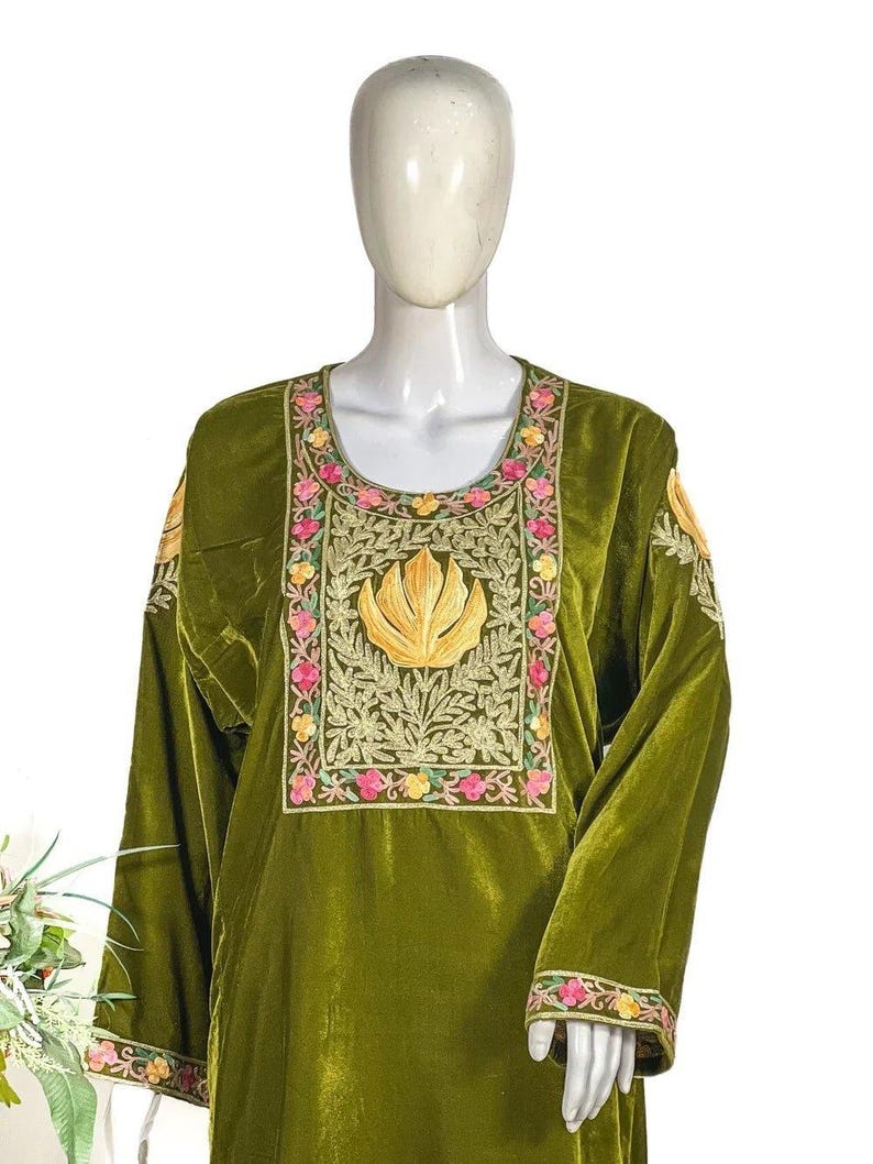 May include: An olive green velvet tunic featuring a scoop neckline and long sleeves. The chest area is adorned with intricate gold, pink, and yellow floral embroidery surrounding a central golden leaf motif. Similar floral embroidery accents the cuffs.