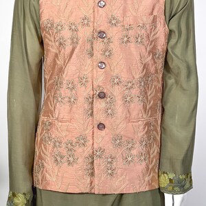 Op de afbeelding: Een perzikkleurig vest met een bloemenpatroon en knoopsluiting. Het vest wordt gedragen over een groen shirt met geborduurde details.