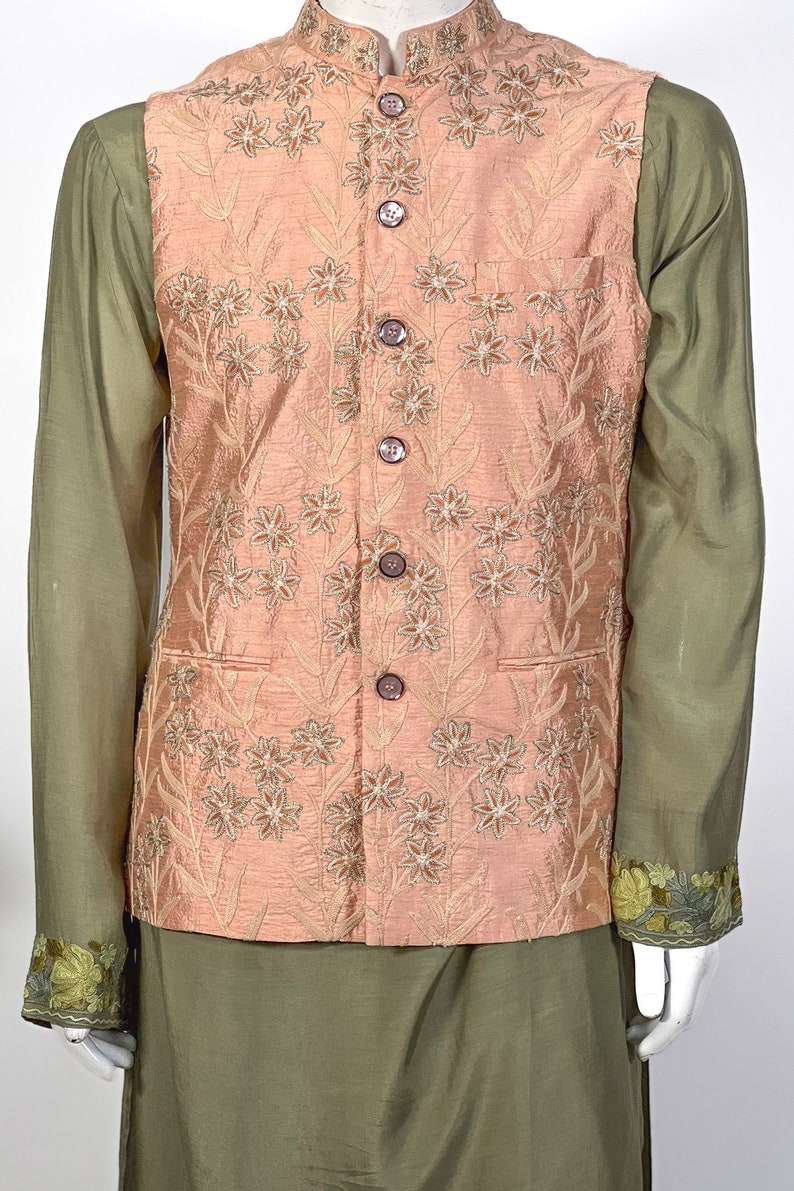 Op de afbeelding: Een perzikkleurig vest met een bloemenpatroon en knoopsluiting. Het vest wordt gedragen over een groen shirt met geborduurde details.