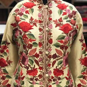 K&ouml;nnte beinhalten: Eine cremefarbene Jacke mit aufwendiger Blumenstickerei in Rot-, Rosa- und Gr&uuml;nt&ouml;nen. Die Jacke hat einen Stehkragen und einen Knopfverschluss.