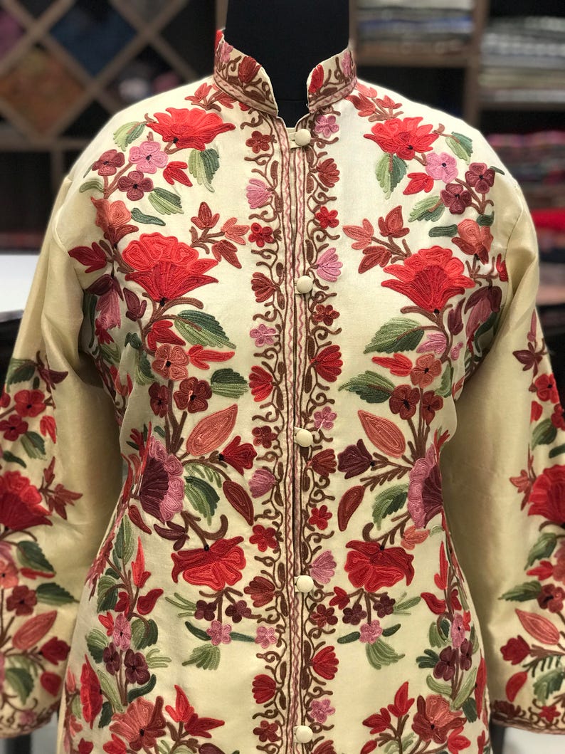 K&ouml;nnte beinhalten: Eine cremefarbene Jacke mit aufwendiger Blumenstickerei in Rot-, Rosa- und Gr&uuml;nt&ouml;nen. Die Jacke hat einen Stehkragen und einen Knopfverschluss.
