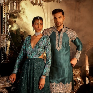 Pode incluir: Um casal vestindo roupas combinando em verde-azulado com bordados intrincados. O vestido da mulher tem um decote profundo e um corpete justo. O traje do homem &eacute; uma kurta de manga comprida com gola mandarim. O casal est&aacute; em p&eacute; em frente a um espelho ornamentado dourado.