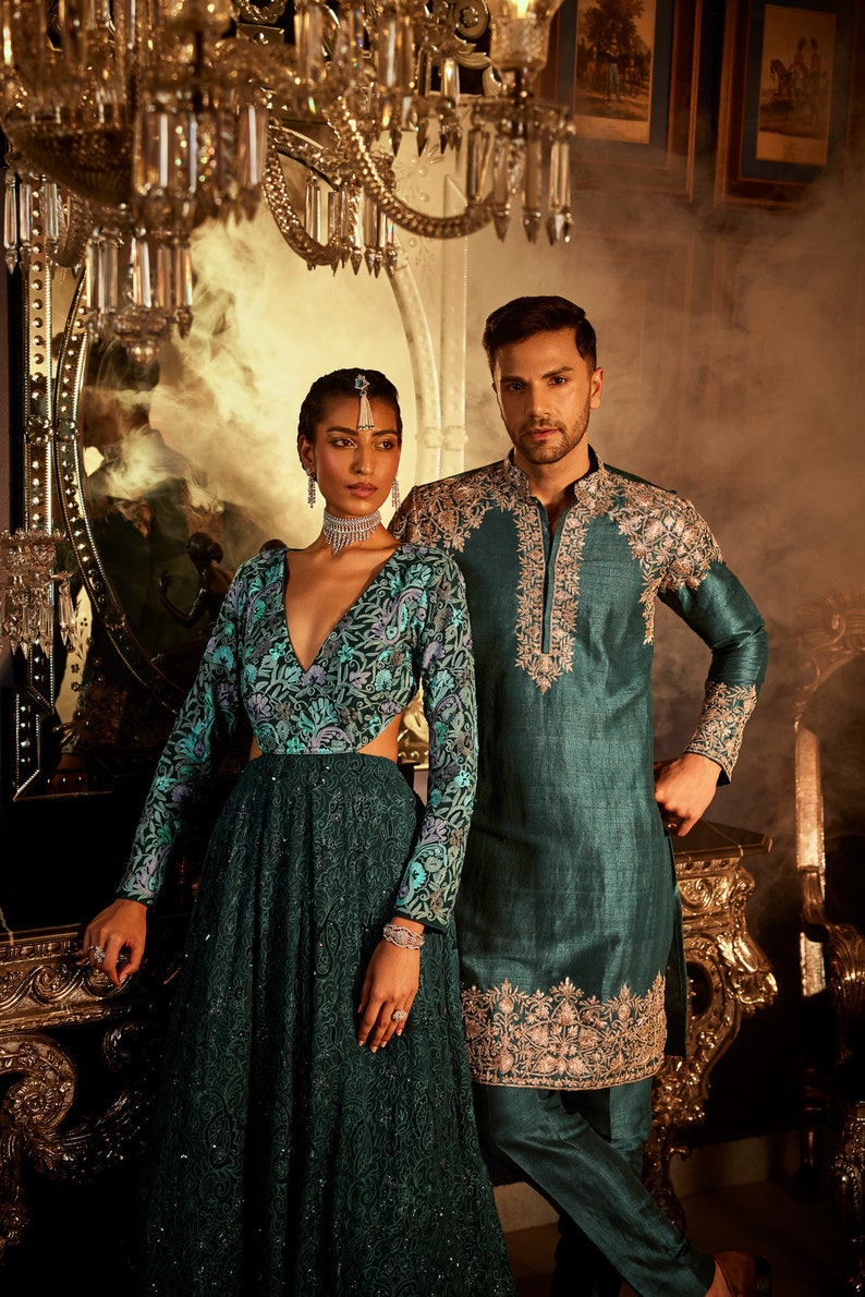 Pode incluir: Um casal vestindo roupas combinando em verde-azulado com bordados intrincados. O vestido da mulher tem um decote profundo e um corpete justo. O traje do homem &eacute; uma kurta de manga comprida com gola mandarim. O casal est&aacute; em p&eacute; em frente a um espelho ornamentado dourado.