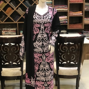 May include: A black long-sleeved tunic featuring a prominent V-neckline and intricate light pink and white floral embroidery covering the front and sleeves. The garment is displayed on a mannequin and includes a matching black shawl draped over one shoulder.