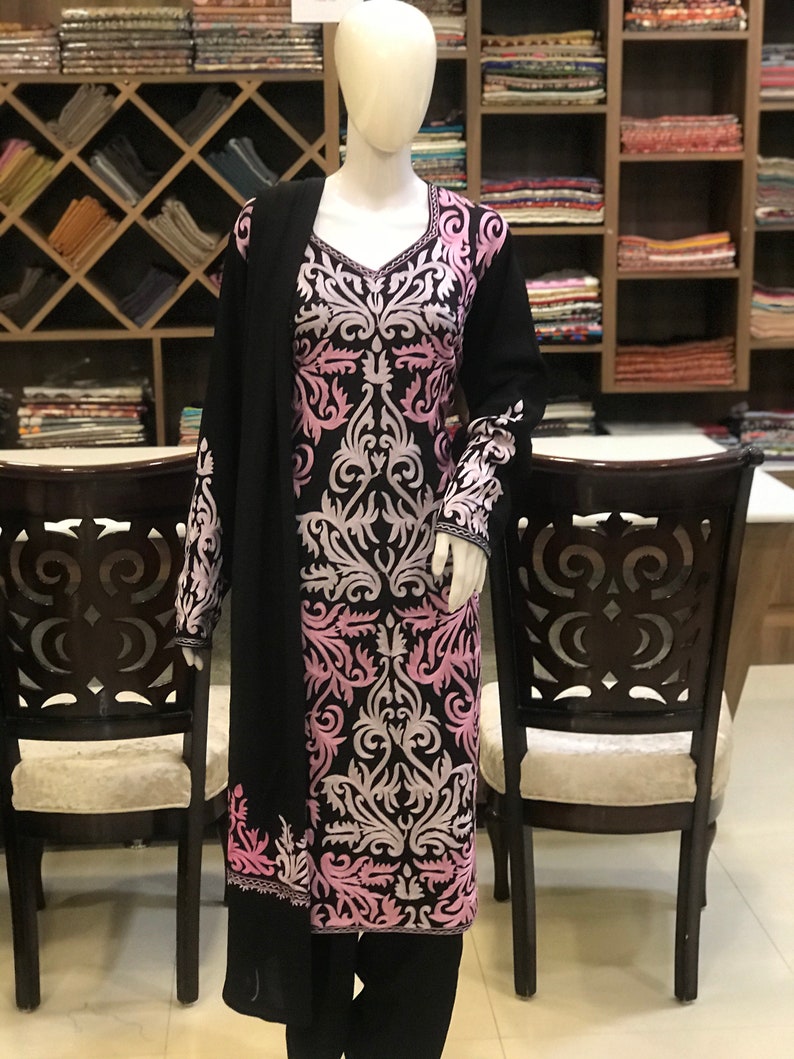 May include: A black long-sleeved tunic featuring a prominent V-neckline and intricate light pink and white floral embroidery covering the front and sleeves. The garment is displayed on a mannequin and includes a matching black shawl draped over one shoulder.