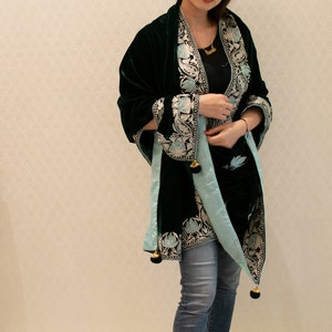 May include: A woman wearing a dark green velvet shawl with light blue and silver embroidery. The shawl has tassels and is worn over a black top and blue jeans.