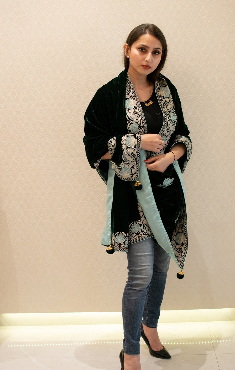 May include: A woman wearing a dark green velvet shawl with light blue and silver embroidery. The shawl has tassels and is worn over a black top and blue jeans.