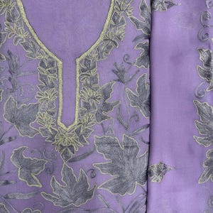 May include: A light purple fabric garment featuring intricate dark purple leaf embroidery and a gold-toned metallic trim around the neckline. The material has a sheer, lightweight texture with a repeating botanical pattern across the surface.