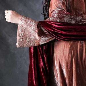 May include: A long-sleeved garment made of dusty rose velvet featuring wide cuffs with intricate gold metallic embroidery. A deep burgundy velvet shawl is draped over the shoulders, adding a rich contrast to the lighter rose-colored fabric.