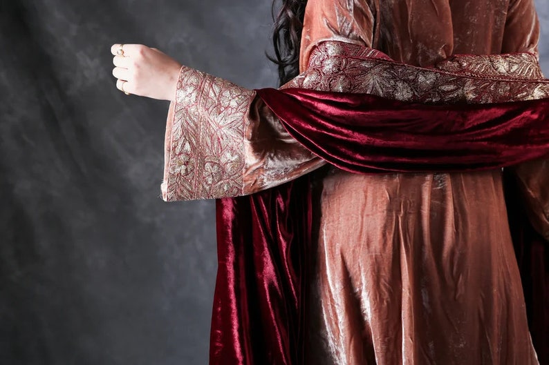 May include: A long-sleeved garment made of dusty rose velvet featuring wide cuffs with intricate gold metallic embroidery. A deep burgundy velvet shawl is draped over the shoulders, adding a rich contrast to the lighter rose-colored fabric.