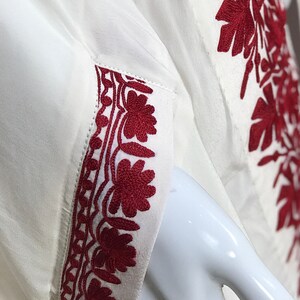 May include: White long-sleeved shirt with red embroidered floral designs on the sleeves and front. The embroidery is detailed and intricate.