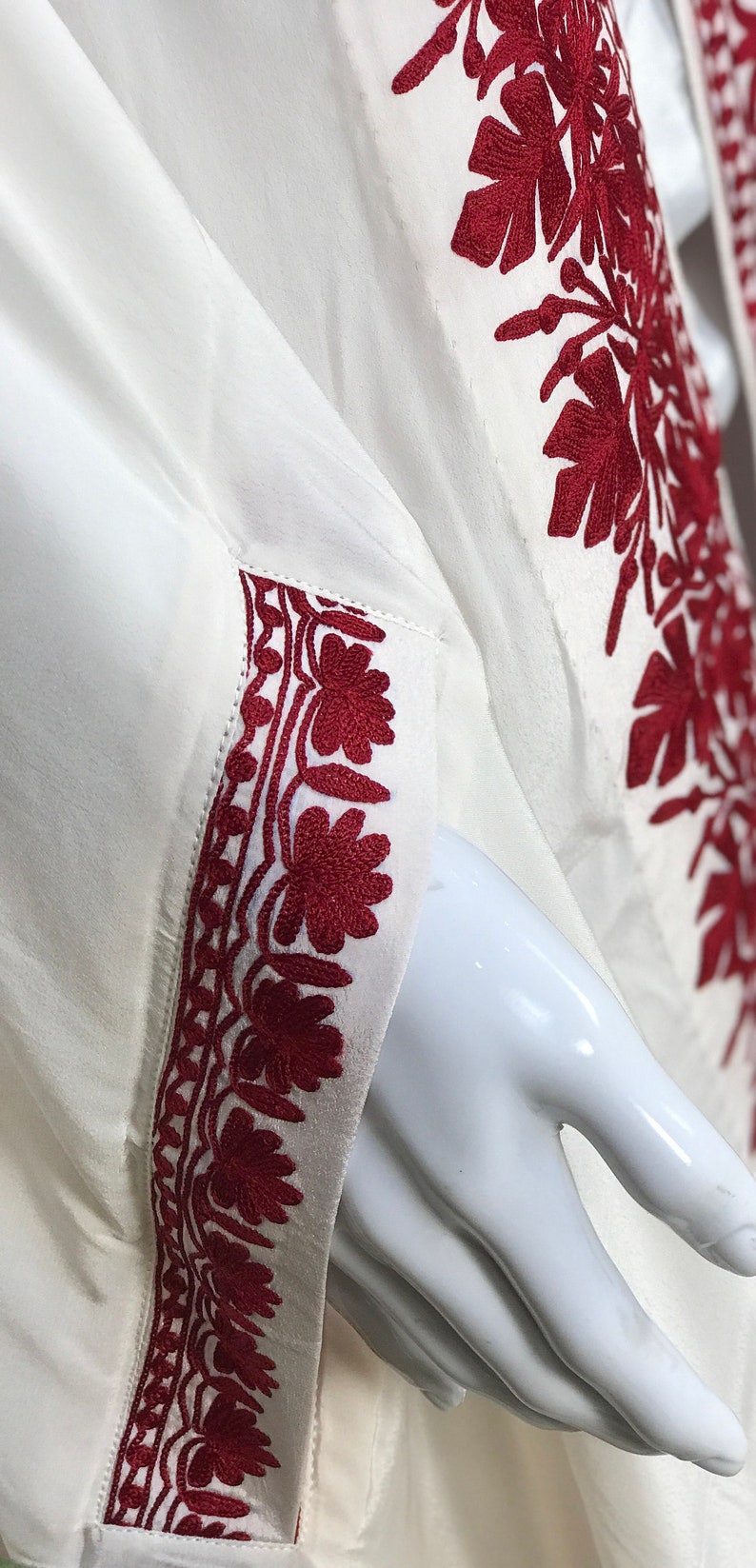 May include: White long-sleeved shirt with red embroidered floral designs on the sleeves and front. The embroidery is detailed and intricate.
