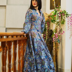 May include: A woman wearing a long, flowing blue and pink floral dress with a V-neckline. The dress has a gradient effect, fading from pink to blue. The woman is standing on a wooden staircase, holding onto the railing.
