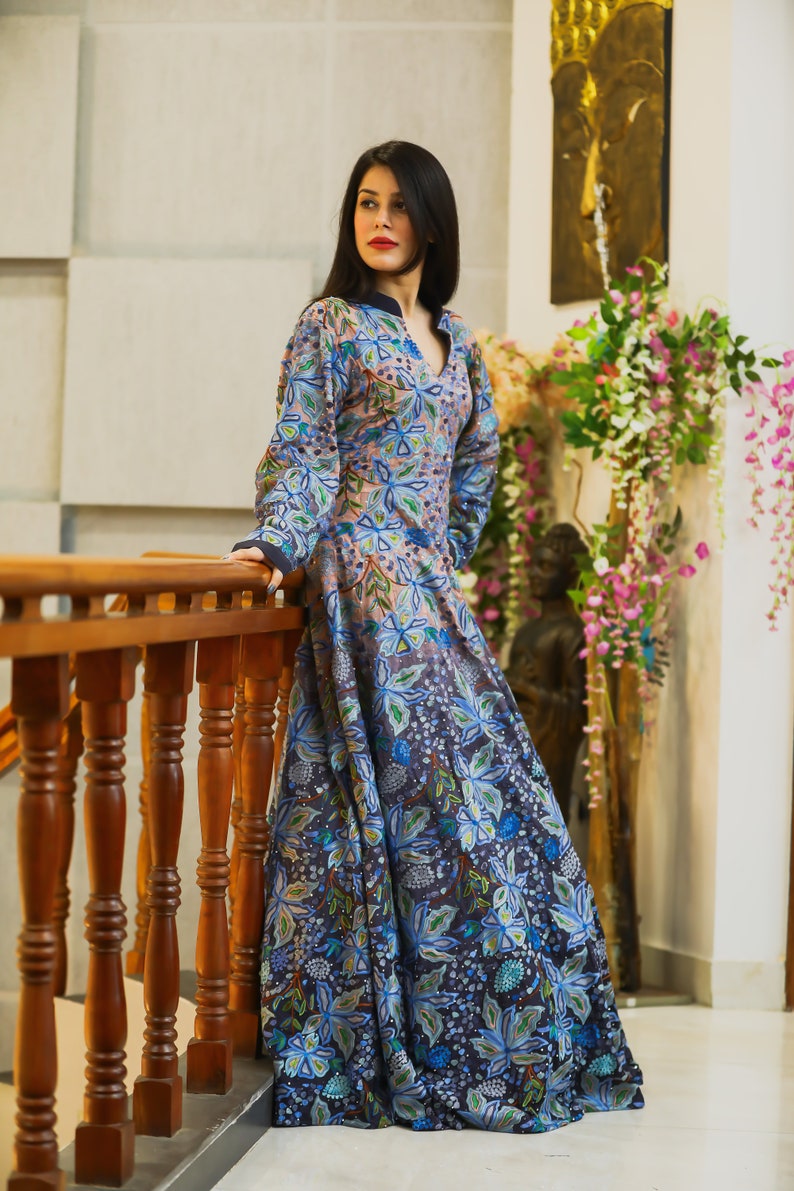 May include: A woman wearing a long, flowing blue and pink floral dress with a V-neckline. The dress has a gradient effect, fading from pink to blue. The woman is standing on a wooden staircase, holding onto the railing.