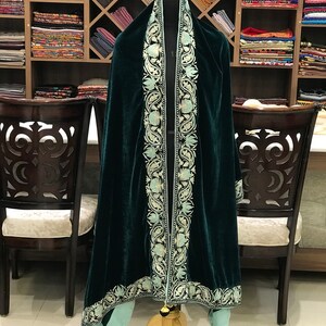 May include: A dark green velvet shawl with a light blue embroidered border. The shawl is draped over a mannequin and has a fringed edge.