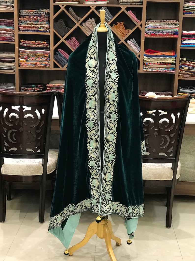 May include: A dark green velvet shawl with a light blue embroidered border. The shawl is draped over a mannequin and has a fringed edge.