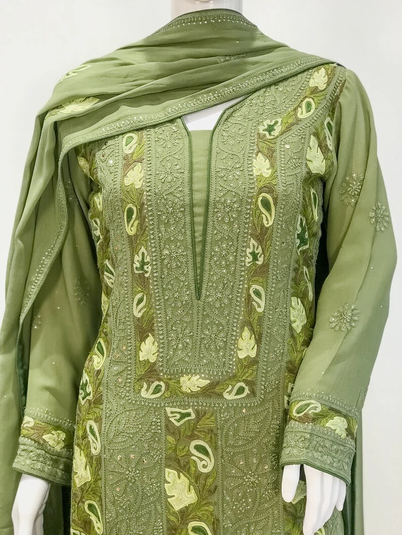 May include: A sage green long-sleeved tunic featuring an intricate embroidered neckline and sleeve cuffs with light green leaf and paisley motifs. The garment includes a matching draped scarf, showcasing detailed needlework and tonal stitching.