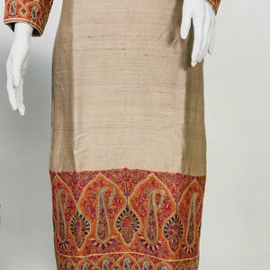 May include: A beige-colored tunic and pants set. The tunic features intricate embroidery in shades of red, orange, and blue along the sleeves and hemline. The fabric has a subtle sheen, suggesting a silk or similar material.