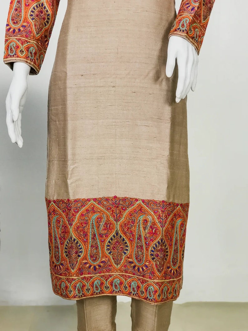 May include: A beige-colored tunic and pants set. The tunic features intricate embroidery in shades of red, orange, and blue along the sleeves and hemline. The fabric has a subtle sheen, suggesting a silk or similar material.