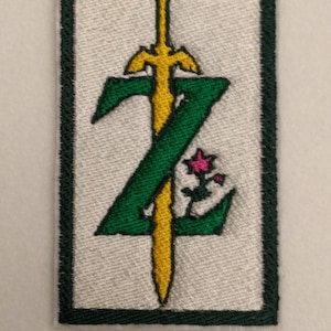 Peut inclure: Patch brodé vert et jaune avec une épée et une fleur, entouré d'une bordure vert foncé. L'épée est en forme de lettre Z.