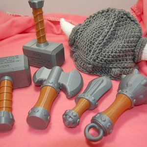 May include: A set of five toy weapons, including a hammer, axe, sword, and mace, all with brown handles and silver-coloured heads. A grey knitted Viking hat with white horns is also included.