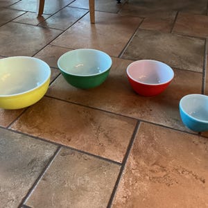 Puede incluir: Cuatro cuencos para mezclar de colores amarillo, verde, rojo y azul. Cada cuenco tiene un interior blanco. Los cuencos están dispuestos sobre una superficie de baldosas marrones. Los cuencos varían en tamaño, siendo el cuenco amarillo el más grande.