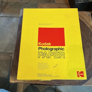 Puede incluir: Una caja amarilla de papel fotográfico Kodak con un cuadrado rojo y texto negro. La caja tiene el logotipo de Kodak y el texto "Made in U.S.A."