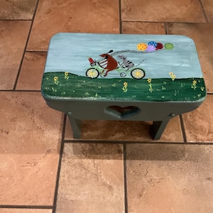 May include: A hand-painted wooden step stool with a whimsical design. The top features a light blue sky with a brown bear and a gray cat riding a bicycle, pulling colorful balloons. The stool has a green base with a heart-shaped cutout.
