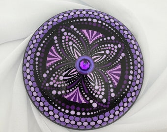 Pode incluir: Um mandala redondo, pintado em roxo e preto em uma pedra, com um strass roxo no centro.