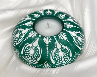 May include: A teal green candle holder with a white mandala design. The holder has a round shape and a small opening in the centre for a tea light candle.
