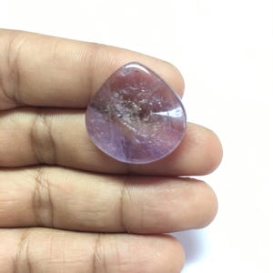 Puede incluir: Un cabuj&oacute;n de piedra preciosa amatista en forma de l&aacute;grima con un tono p&uacute;rpura claro y una apariencia transl&uacute;cida.