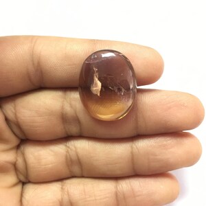 Puede incluir: Una piedra preciosa ovalada lisa con un degradado de color de marr&oacute;n a &aacute;mbar. La piedra es transl&uacute;cida con inclusiones internas visibles.