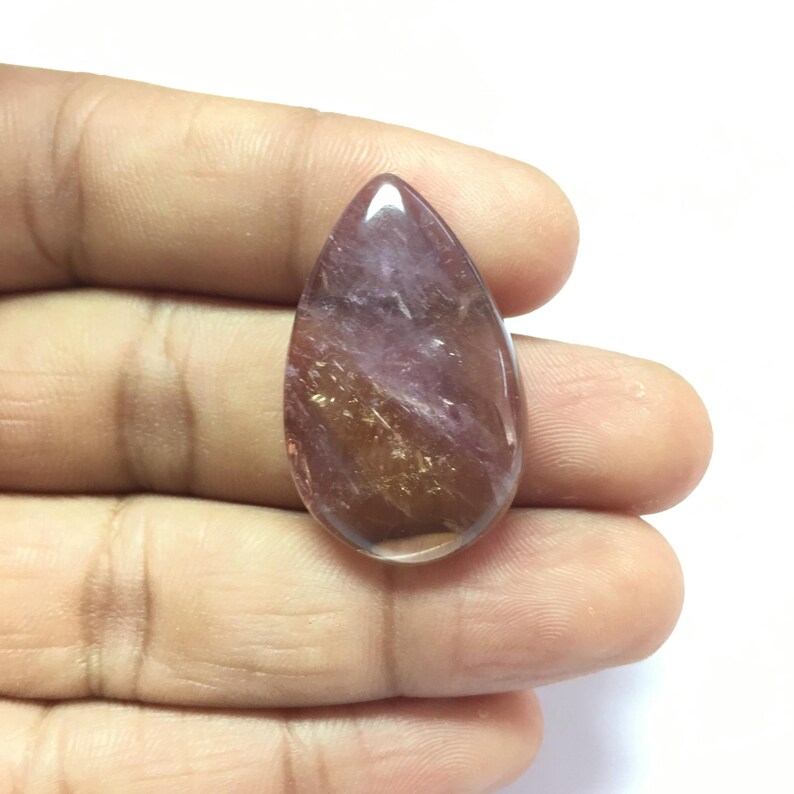Puede incluir: Una piedra preciosa transl&uacute;cida en forma de l&aacute;grima con un color rosa y dorado. La piedra preciosa tiene una superficie lisa y pulida.