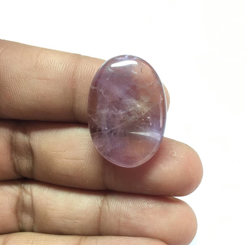 Puede incluir: Una piedra preciosa ovalada, lisa, de color p&uacute;rpura y blanco, con una apariencia transl&uacute;cida.