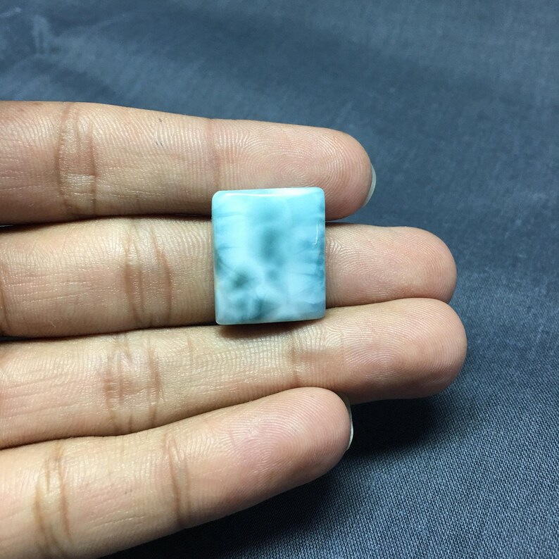 May include: A square-shaped, light blue gemstone with white and gray swirls. The gemstone is a cabochon cut.