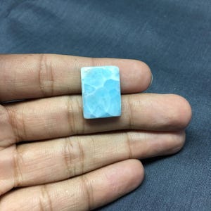 May include: A light blue cabochon gemstone with a cloudy, marbled pattern. The gemstone is rectangular in shape and has a smooth, polished surface.