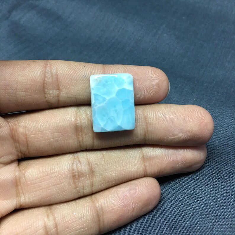 May include: A light blue cabochon gemstone with a cloudy, marbled pattern. The gemstone is rectangular in shape and has a smooth, polished surface.
