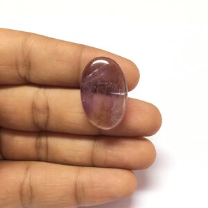Puede incluir: Una piedra preciosa ovalada lisa de color p&uacute;rpura con un tono rosa claro. La piedra preciosa es transl&uacute;cida y tiene un ligero brillo.