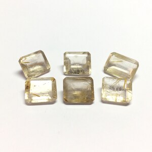 Pode incluir: Seis pedras preciosas quadradas, facetadas e transparentes com inclusões douradas. As pedras preciosas estão dispostas em semicírculo sobre um fundo branco.