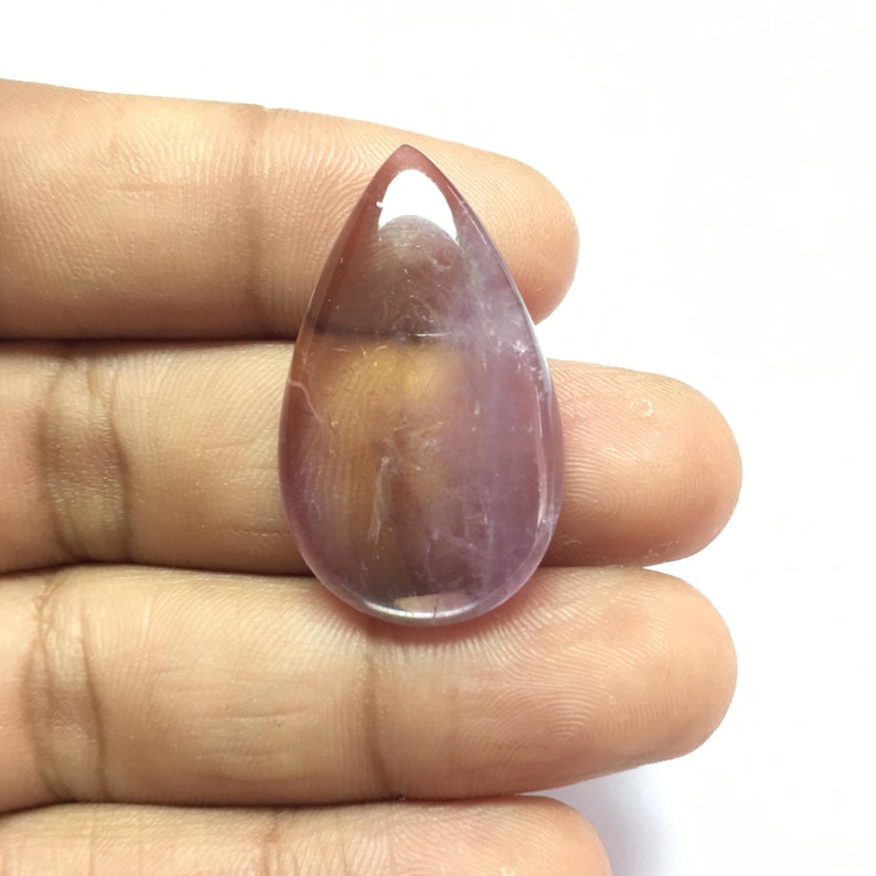 Puede incluir: Una piedra preciosa en cabuj&oacute;n en forma de l&aacute;grima con un degradado de color p&uacute;rpura y marr&oacute;n. La piedra preciosa es lisa y pulida.